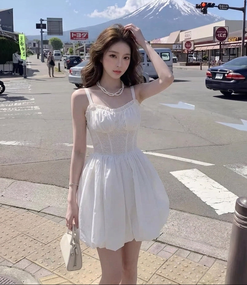 A woman stands on a city street wearing a white sleeveless mini dress with a gathered bodice and flared skirt. A snow-capped mountain is visible in the background under a blue sky, with cars and storefronts around her.