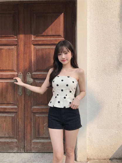 A woman stands in front of a rustic wooden door and textured wall, wearing a strapless white tube top with black polka dots and black shorts.