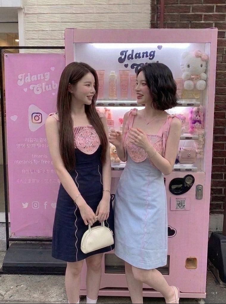 Two women standing in front of a pink vending machine and sign in front of a brick building. One woman is wearing a navy blue dress with a pink lace bodice and holding a cream handbag.