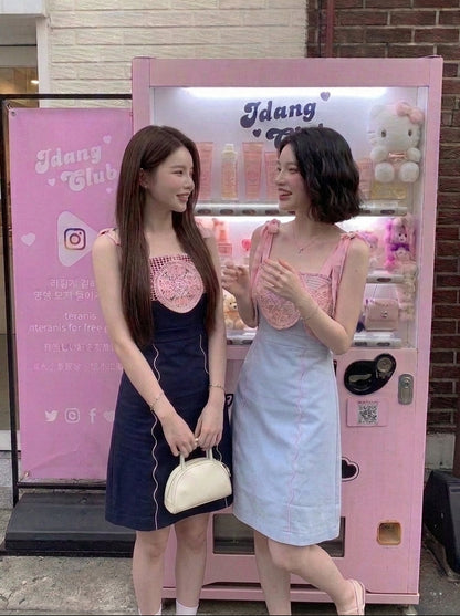 Two women standing in front of a pink vending machine and sign in front of a brick building. One woman is wearing a navy blue dress with a pink lace bodice and holding a cream handbag.